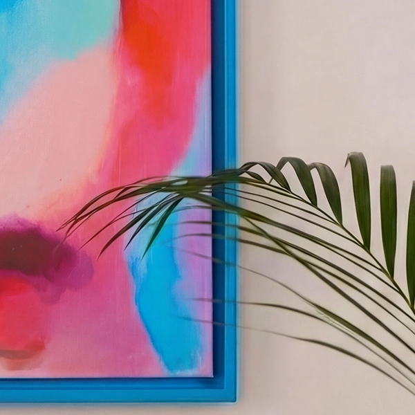 Marco azul con una imagen colorida en rosa y turquesa en una pared beige claro. Una hoja de palma verde sobresale en la imagen.